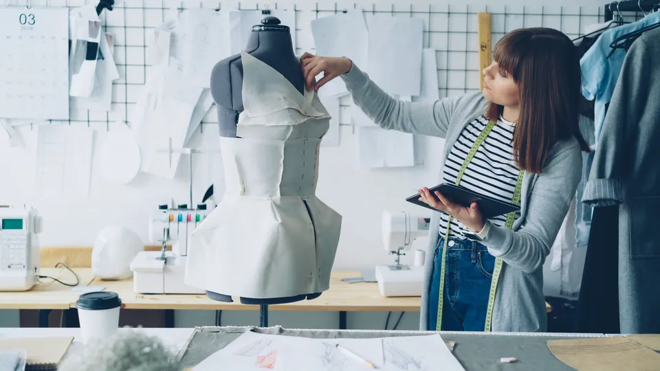 Designer working on a dress on a mannequin in a bright studio, with paper patterns on the wall