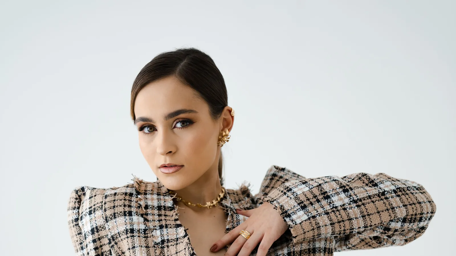 Model in a matching check blazer and skirt with a gold necklace, seated in a white studio
